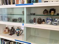 Two shelves showing assorted small pottery items: dark vases, small bowl, red vase, rustic jar, blue and white porcelain jars and bowl, fish motif vase, colorful teapot