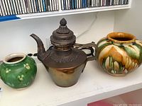 Three items on shelf: small green vase, ornate metal teapot, large multicolor drip-glaze vase