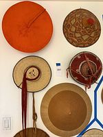 Photo showing six woven hats displayed on a white wall, highlighting different styles and colors, including the orange spiral hat and conical patterned hat.