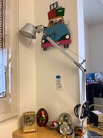 Photo showing metal adjustable desk lamp on a wooden cabinet alongside a brass-colored clock, several decorative eggs, small globe paperweights, and a miniature soccer ball figurine, with a colorful wall clock shaped like a car with passengers in the background.