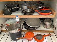 Two-shelf overview showing All-Clad nonstick pots, aluminum pans and Copco orange cast iron cookware