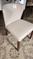 Fabric-covered side chair with beige upholstery and wooden legs on patterned floor.