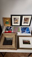 Overview photo showing 2 framed art prints in black frames, 1 gold framed oil painting of flowers, 3 colorful abstract acrylic paintings without frames laid on table, and 1 framed square mirror with carved wooden frame.