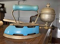 Photo of antique blue metal flat iron with brass gas burner attached to the side, showing front and side views.
