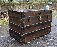 Diagonal view of the antique wooden trunk outdoors, showing metal reinforcements and overall condition.