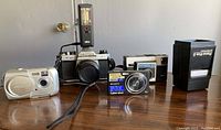 Four cameras and a hand-held slide viewer displayed on a wooden table, including Pentax K1000 with flash, Sony Cyber-shot digital camera, Olympus Camedia D-395, Instamatic 126 film camera, external zoom lens and flash, plus cords.
