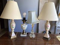 Four lamps on table: two rosebud floral lamps with cream fabric shades, one rosebud floral lamp with cream shade on left, one plastic crystal-style lamp with pale blue velvet shade in center