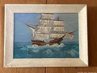Framed oil painting showing a large sailing ship in full sail on the ocean waves, framed in a whitewashed wood frame.