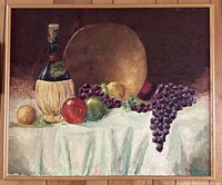Framed still life painting showing fruit including grapes, apples, lemon, and a straw-covered bottle on a white draped cloth with a large round golden background object.