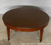 Photo shows a round Mid-Century Modern coffee table with a wood finish top and tapered wooden legs with casters, placed on a concrete floor.
