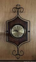 Photo of a mid-century modern electric wall clock mounted on a wooden wall. Clock features a gold-tone round face with Roman numerals, set in a wooden backplate and surrounded by decorative black metal scrollwork.