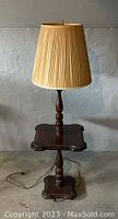 Full view of one vintage floor lamp and end table combo. Shows pleated beige lamp shade, dark wood turned column, square small table surface and base.