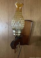Single amber glass hurricane lamp electric wall sconce mounted on wood base shown against wood paneled wall.