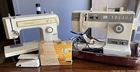 Two cream-colored Kenmore sewing machines on a wooden surface, one with attached cords, buttonholer, and manual booklet.