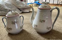 Photo showing a vintage ceramic chocolate set including a large lidded pot with spout and handle alongside a smaller covered sugar bowl, both decorated with floral patterns and pink trim.