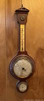 Full view of the vintage wooden weather station hanging on a wood-paneled wall, showing thermometer at center, large barometer dial below, and smaller hygrometer dial at bottom.