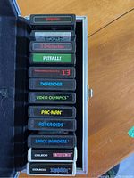 Opened silver metal carrying case showing 12 Atari-era game cartridges in foam slots