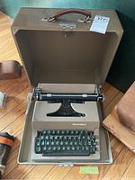 Manual typewriter in open brown carrying case. Beige body with dark green keys.