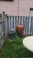 Wide view showing garden wheelbarrow, hose reel, and garden hose against wooden fence on grassy patch.