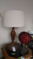 Photo of a carved wood base lamp with a white pleated fabric drum shade, placed on a wooden table.