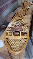 Photo showing close-up of one wooden snowshoe with leather bindings and rawhide webbing on a wooden surface.