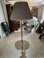 Full view of the lamp with grayish pleated fabric shade and brass finish metal with attached glass top table surface.