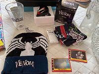 Photo showing the Venom knit hat, Venom glass, mini planter with original box, scarf, and two Alien collector cards on a white surface.