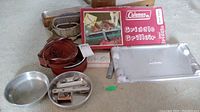 Overview photo showing Coleman Griddle box, metal griddle plate, camping dishes including frying pan and plate, red nested dish set, wooden spatula, metal utensils, and match tin.