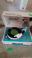Photo showing open vintage record player with Mickey Mouse graphic inside lid, record on turntable, and Mickey glove tonearm in teal and white case.