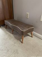 Side and angled view of a brown vinyl cushioned stool with hourglass curved seat on four wooden tapered legs.