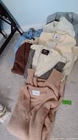 Several wool sweaters in neutral colors stacked together on carpet showing overall lot contents including multiple visible brand labels.