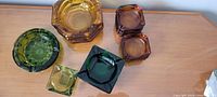 Photo showing a collection of six colored glass ashtrays including green, amber, and dark green varieties arranged on a wooden surface.