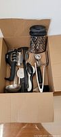 Top-down view in cardboard box showing 13 assorted kitchen utensils including graters, ladles, scoops, whisks and peelers in black and metal finishes.