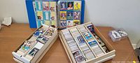 Two white boxes filled with assorted sports cards on a floor, next to a blue binder with sports cards inside, open on a page.