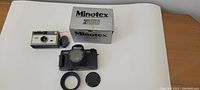 Photo of the Minotex 2000 camera with box and accessories (lens cap and ring) on table, showing front and top details.