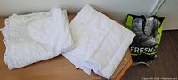 Two folded white banquet table cloths with visible Battenburg lace detail and packaging bag nearby, placed on wooden table and floor visible.