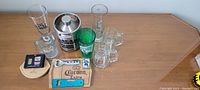 Photo of 12-piece beer enthusiast paraphernalia set on table featuring branded glasses, mugs, can cooler, coasters, bottle opener, and tray.
