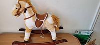 Full front-side view of the plush rocking horse showing the fabric texture, brown saddle, mane, and wooden rockers.