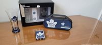 Photo showing the Toronto Maple Leafs branded bar set including a pilsner glass, whiskey glass with bar tools in original box, and unopened collector cards on a table.