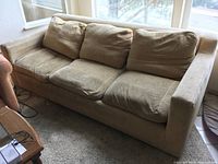 Tan down-filled couch with three seat and back cushions shown from an angle. Visible wear and some dirt marks on cushions. Wooden frame and straight armrests in good condition.