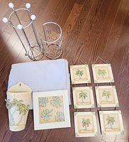 Photo showing two metal toilet paper holders, ceramic wall pocket, light blue towel, framed leaf print, and six small palm tree plaques on wood floor