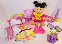 Full view of Cabbage Patch Kid doll with hair in pigtails, seated on white surface surrounded by all accessories including clothing, towels, rubber duck, and pink swim fins.