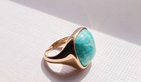 Close-up photo of ring showing large blue-green cabochon stone and gold-tone band on white background under natural light.