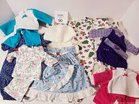 Lot of assorted doll clothing including dresses, jackets, pants, hats, and tops shown on a white background.