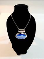 Necklace displayed on a black bust form showing front view of chain with oval blue pendant in silver-tone metal frame.