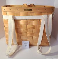 Front view of rectangular woven wood basket with white shoulder straps attached and visible wooden handle on top edge.