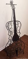 Full front side view of the black metal cello shaped wine rack standing upright on wooden floor against wall.