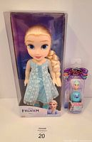 Front view of boxed Disney Frozen Elsa doll approximately 11 inches tall wearing a turquoise dress with snowflake pattern next to a Hatchimals Mini Pixies collectible toy in packaging
