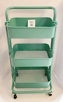 Front view of mint green utility rolling cart showing three tiers: solid bin on top, two mesh trays below and four black swivel castors.