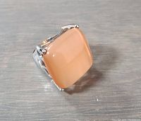 Close-up photo of ring showing square polished peach-colored stone set in stainless steel band on a grey background.